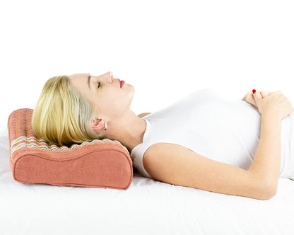 A person with blonde hair rests on their back, eyes closed and hands on their stomach, using the pink TAO-Mat® Pillow Soft InfraMat Pro® by HealthyLine for neck support. They wear a white sleeveless top and lie peacefully against a plain white background.