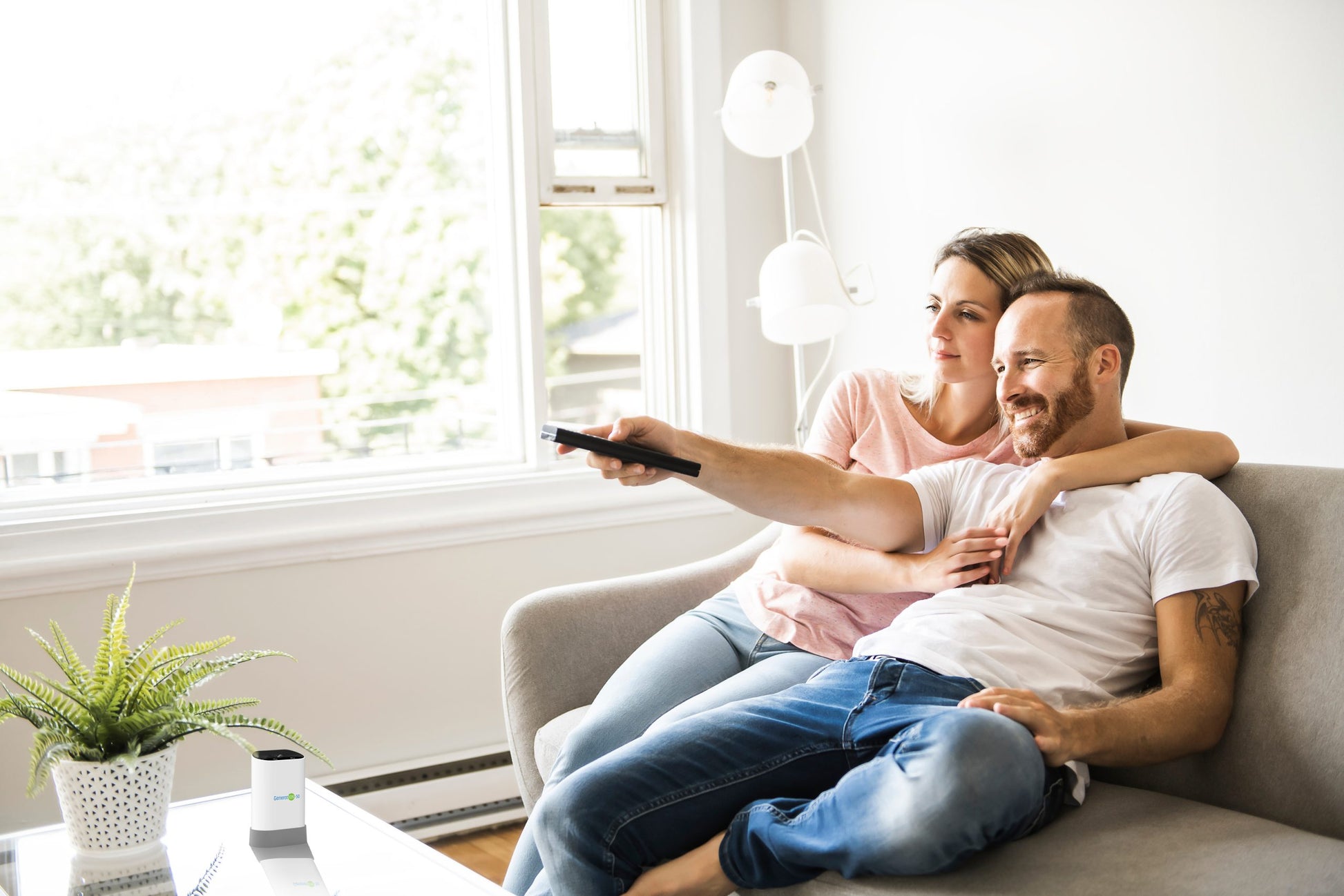 A couple relaxes on a couch in a sunlit living room. The man holds a TV remote, both smiling as they gaze ahead. A potted plant adorns the coffee table, complemented by the Generation-50 Air Purifying Device from HealthyLine, while sunlight pours through the large window.