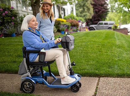 An older woman in a blue sweater smiles while riding her Journey Health & Life Style So Lite Scooter along a sidewalk with a younger companion, enjoying the scooter’s lightweight mobility near a green lawn and colorful flowers.