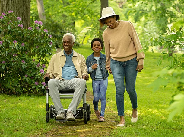 A man in a Zoomer wheelchair from Journey Health & Lifestyle, steered by a joystick, is guided along a garden path with green trees and flowers. A child holds the wheelchair, while a smiling woman wearing a hat walks beside them.