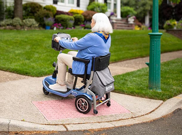 An older adult with white hair and a blue jacket rides a Journey Health & Life Style So Lite Scooter on a suburban sidewalk ramp, enjoying its lightweight design and advanced safety features.