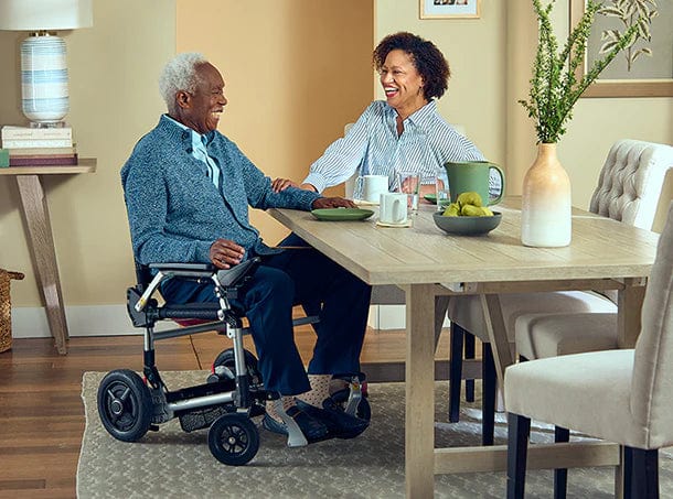 A man in a Journey Health & Life Style Zoomer (w/ Joystick) wheelchair smiles as he navigates with one hand, holding hands with a woman at the warmly decorated dining table. Surrounded by plants and books, they appear happy and engaged in conversation.