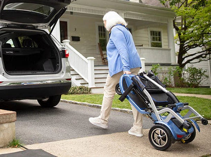 An older adult with white hair, in a blue sweater and beige pants, carries a Journey Health & Life Style So Lite Scooter toward the open trunk of a car parked in front of a house with white siding and a porch.