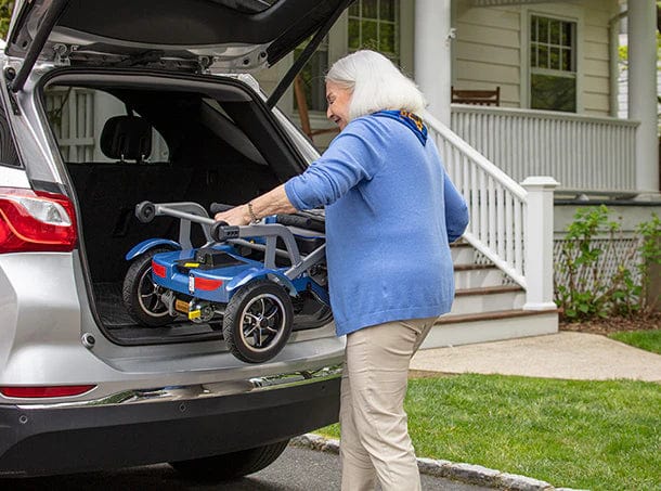 An older woman with gray hair loads a folded, lightweight Journey Health & Life Style So Lite Scooter into the trunk of an SUV parked outside a house with a porch.