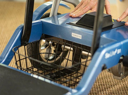 Close-up of hands pressing a button on the Journey Health & Life Style So Lite Scooter, highlighting its front basket and wheels on a woven brown floor mat—showcasing the scooter’s lightweight mobility and practical design.