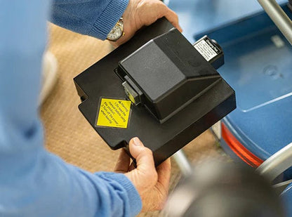 A person in a blue sweater holds a black battery with a yellow warning label beside the Journey Health & Life Style So Lite Scooter, showcasing its lightweight mobility and safety-focused design.