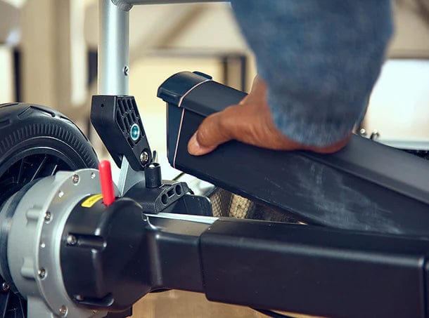 A person is skillfully inserting a removable battery pack into a Zoomer (w/ Joystick) by Journey Health & Lifestyle. The scooter's wheel, part of its frame, and a red lever are visible indoors, highlighting the precise assembly process.
