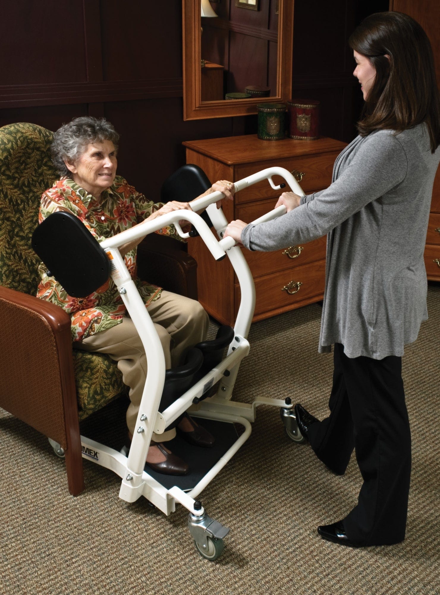 In a warmly lit room with wooden furniture, a caregiver helps an elderly person in a floral armchair using the Graham Field Stand Assist, a mobility aid with wheels.