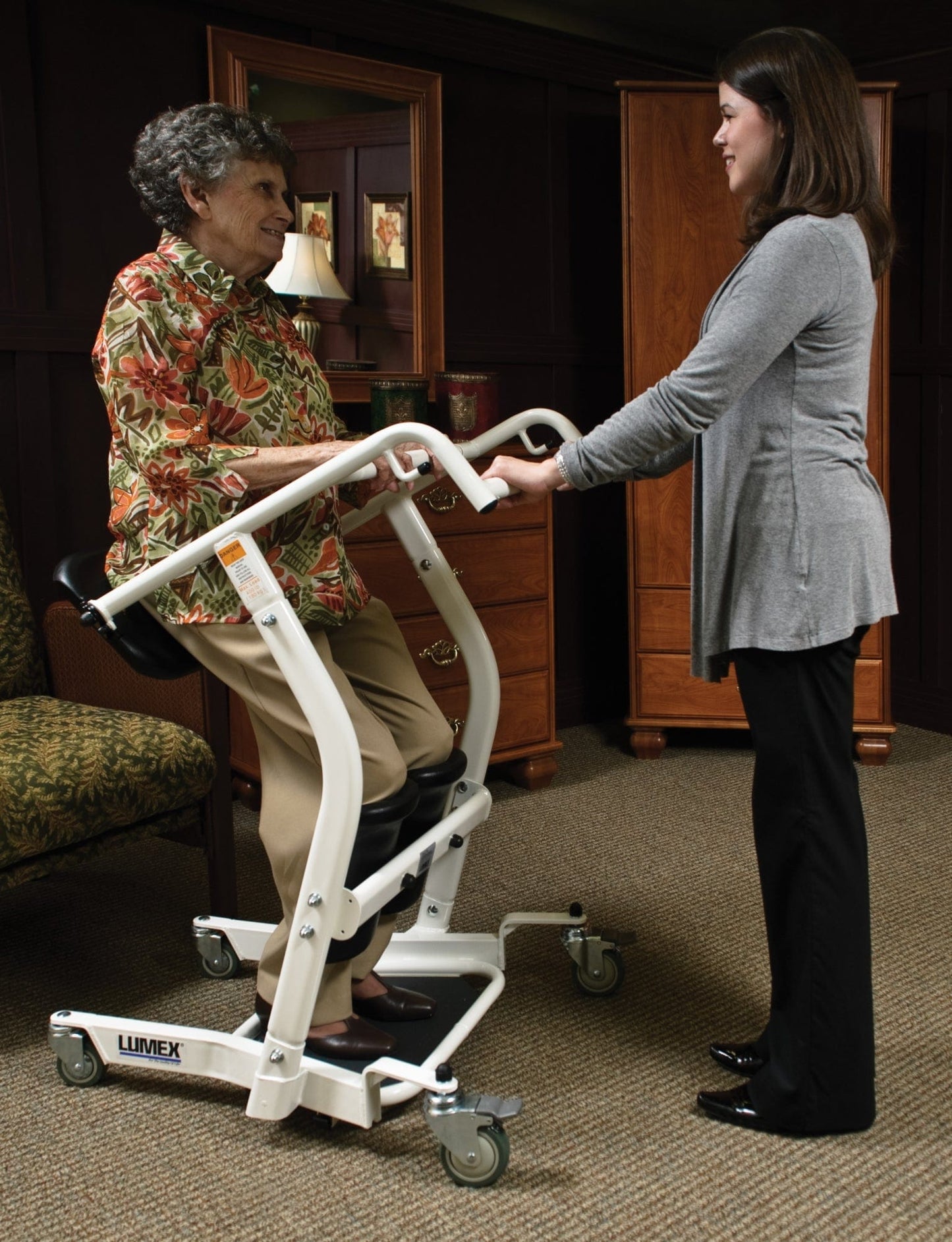 An elderly woman in a floral shirt sits on a Graham Field Stand Assist device. A young woman in a gray top stands before her, smiling warmly, amid dark wood paneling and a green chair, creating a cozy atmosphere.