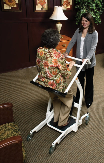 An elderly woman uses a Graham Field Stand Assist while a younger woman helps her in a carpeted room with floral artwork, a lamp, and a nearby chair with floral upholstery.