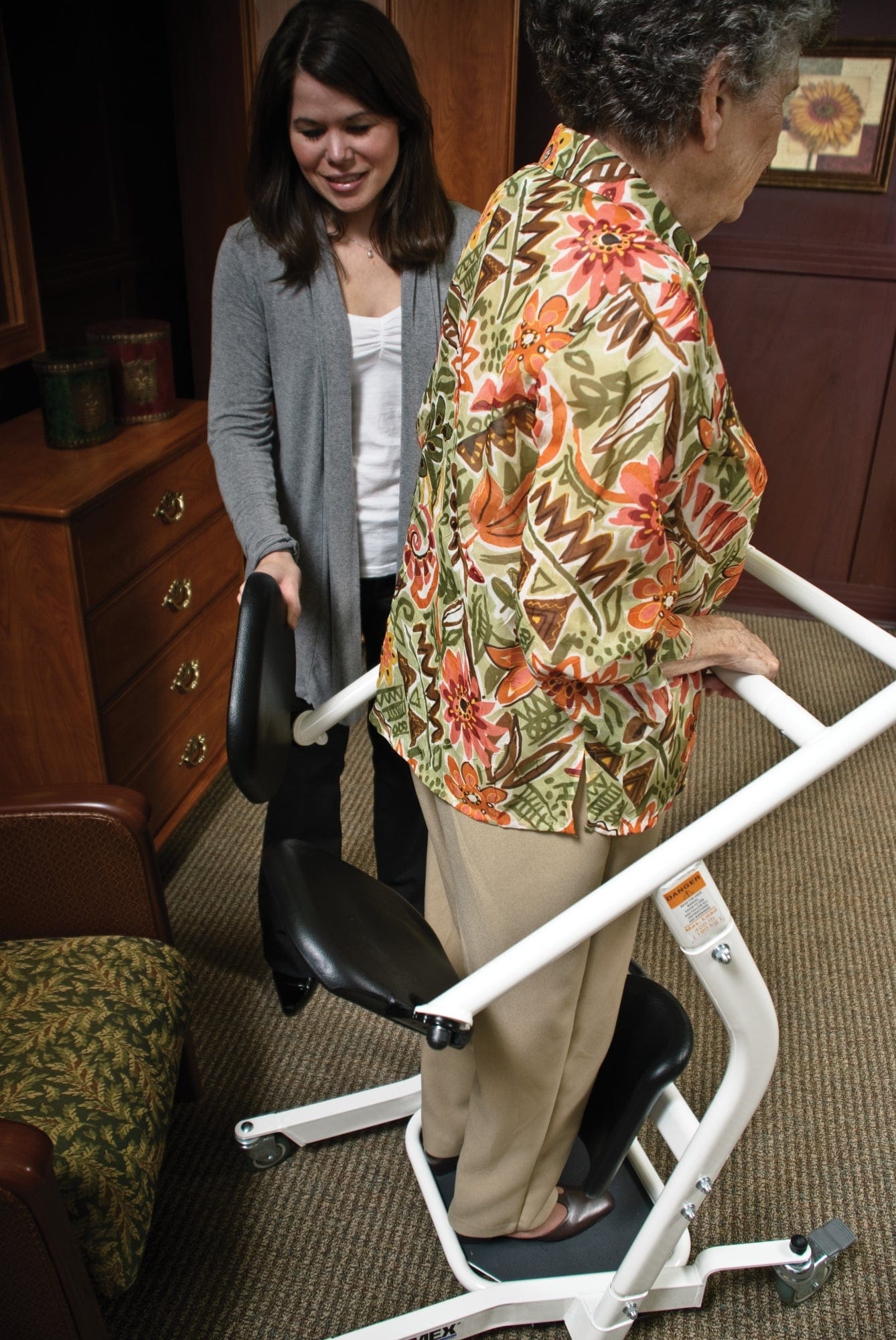 In a room with wooden furniture, a caregiver helps an elderly woman using a Graham Field Stand Assist. The woman wears a colorful floral shirt and beige pants, while the smiling caregiver is in a gray cardigan over a white shirt.