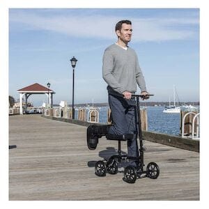 A man uses a Drive Medical Knee Walker Economy Folding RTL BX on a wooden pier by the water, enjoying mobility during recovery with boats and a gazebo in the background under a clear sky.