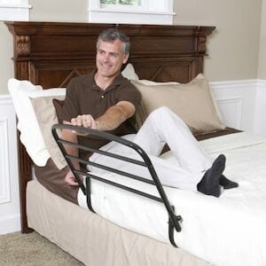 A man sitting on a bed showcases the Stander 30" Safety Bed Rail attached to its side. The room is adorned with beige walls and a wooden headboard. He smiles warmly, wearing a brown shirt and light pants.