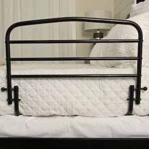 A Stander 30" Safety Bed Rail in black metal is secured to the side of a bed adorned with white quilted bedding, with a lamp and nightstand partially visible in the background.