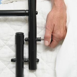 A hand gently rests on a white quilted surface beside the dependable Stander 30" Safety Bed Rail in black metal, ensuring safety with every slumber.